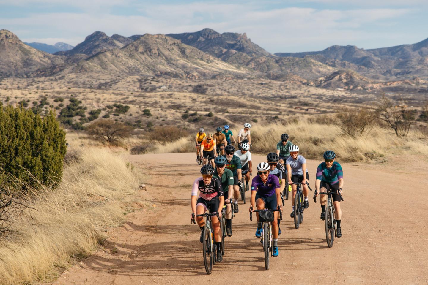 Cyclists riding on a dirt road with mountains in the background.